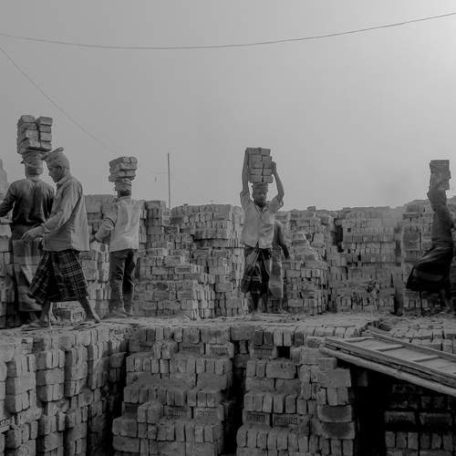 A brickfield worker pushes a handcart loaded with freshly molded clay past towering stacks of sun-dried bricks, as smoke rises from the kiln chimney in the background. In Bangladesh’s brick-making yards, laborers spend long hours shaping, carrying, and fi
