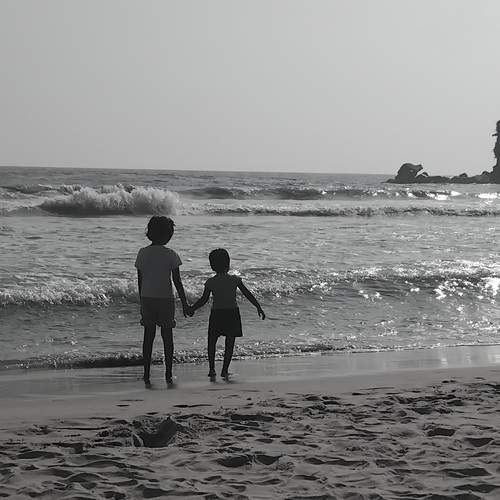 ​A high-contrast, black-and-white shot of two young children standing at the water\\\'s edge.