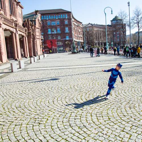 A spectacular wide angle photograph showing a young child in a blue carnival costume running on a massive stone pavement square near historic buildings