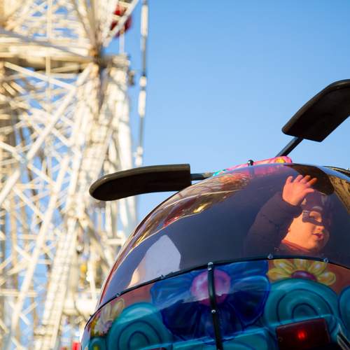 A beautiful low angle photograph focusing on a happy child waving from a colorful helicopter amusement ride cabin with a massive white ferris wheel background