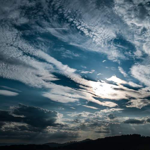 A magnificent high angle aerial drone photograph focusing on a dramatic cloudy blue sky with bright sun rays breaking through texture layers over a dark picea abies spruce forest silhouette