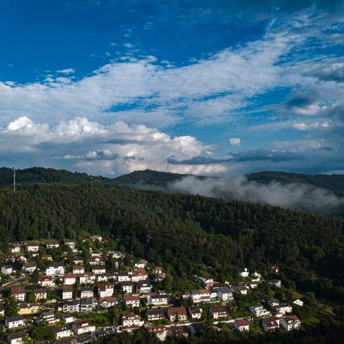 A magnificent high angle aerial drone photograph showing a scenic European village surrounded by dense picea abies spruce forests under a dramatic cloudy blue sky