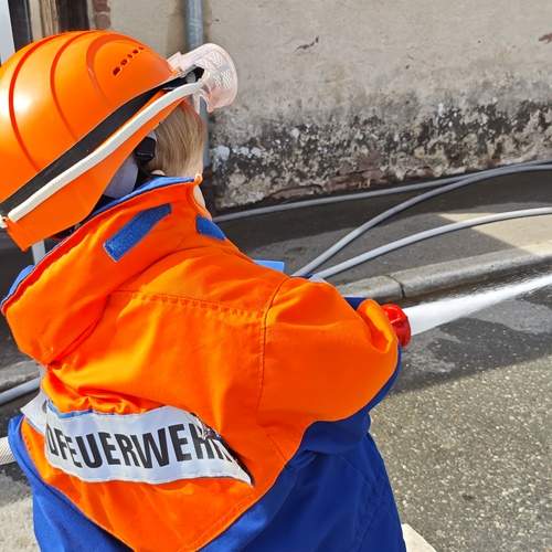 A detailed back angle photograph showing a young boy wearing a bright orange firefighter protective helmet and uniform while spraying water with a hose in a street near an old building wall