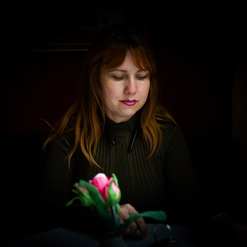 A beautiful detailed close up moody low key portrait photograph showing a thoughtful adult woman sitting inside a dark restaurant next to a pink rose rosa flower during a sunny day