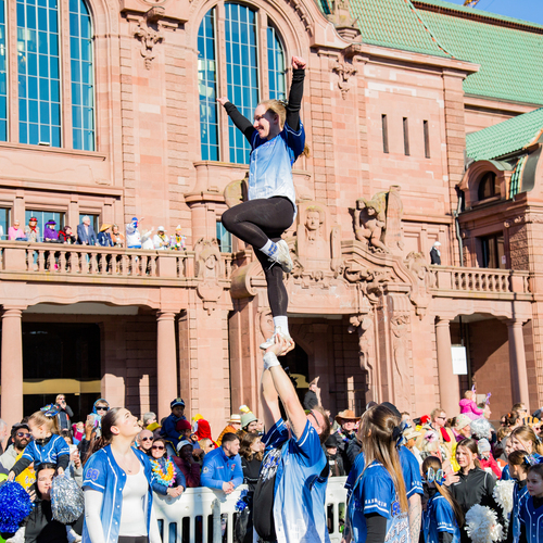 A joyful detailed wide angle action photograph showing cheerleaders performing stunts with many human spectators in front of a historic building during a sunny spring day