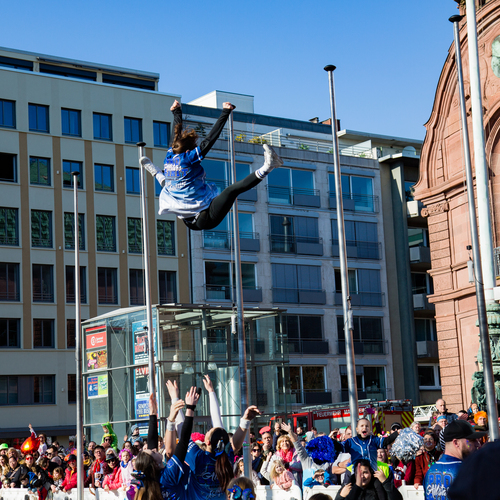 A joyful detailed wide angle action photograph showing cheerleaders performing high aerial acrobatic stunts with many human spectators in front of city buildings during a sunny spring day