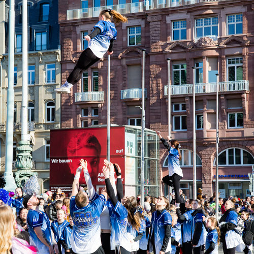 A joyful detailed wide angle action photograph showing young cheerleaders performing high aerial tossed stunts with many human spectators in front of historic buildings during a sunny spring day