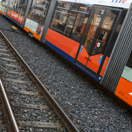 A modern orange commuter tram train moving along steel railway tracks with crushed stone ballast background near small roadside grass lawn during a sunny day