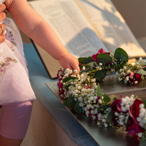 A detailed close up lifestyle photograph showing a little baby hand touching a delicate gypsophila paniculata flower wreath near an open book during a sunny day