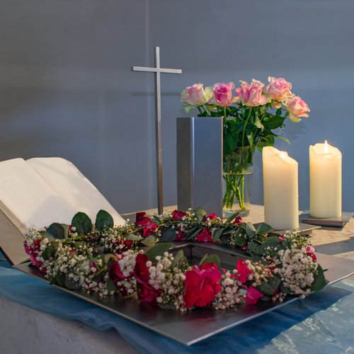 A detailed horizontal interior lifestyle photograph showing a christian church baptismal font holy water vessel with gypsophila wreath open bible cross candles and pink rosa hybrid roses during a sunn