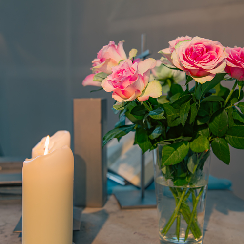 A detailed close up interior lifestyle photograph showing a bouquet of pink rosa hybrid roses in a glass vase with burning white candles inside a church during a sunny day
