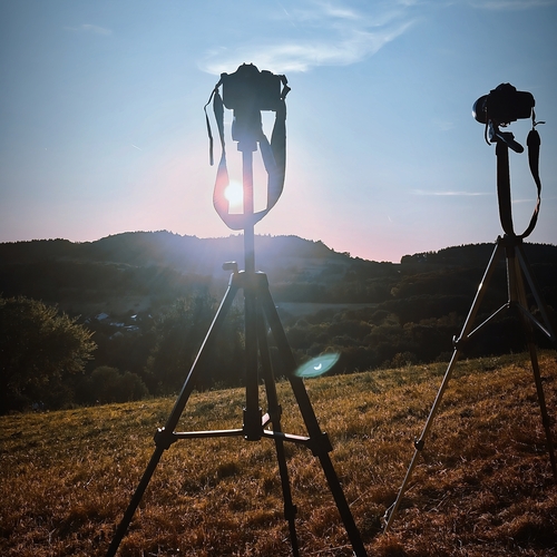 A detailed vertical photograph showing two cameras on tripods shooting sunset landscape silhouette over green hills field
