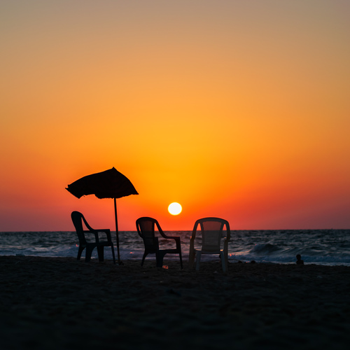 At the end of the day, the beach falls silent beneath a burning sky. Empty chairs and a worn umbrella stand between the fading light and the restless sea, creating a quiet moment where human presence meets the vastness of nature.