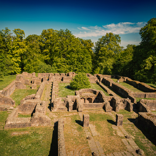 A detailed wide landscape photo showing historic medieval stone ruins of St Michael Monastery surrounded by green forest trees under sunny blue sky