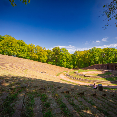 A detailed wide landscape photo showing historic open air amphitheater Heidelberg Thingstaette surrounded by green forest trees under sunny blue sky