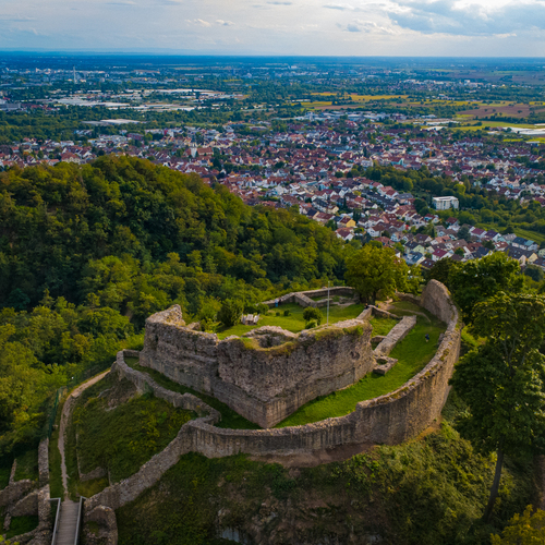 A detailed aerial drone landscape photo showing ancient stone ruins of Strahlenburg Castle on forested mountain ridge overlooking European town valley